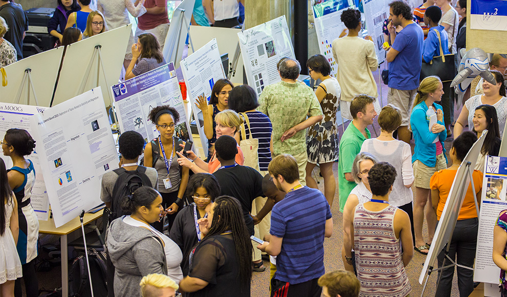 Students, faculty, and visitors gather around posters presenting student summer research.