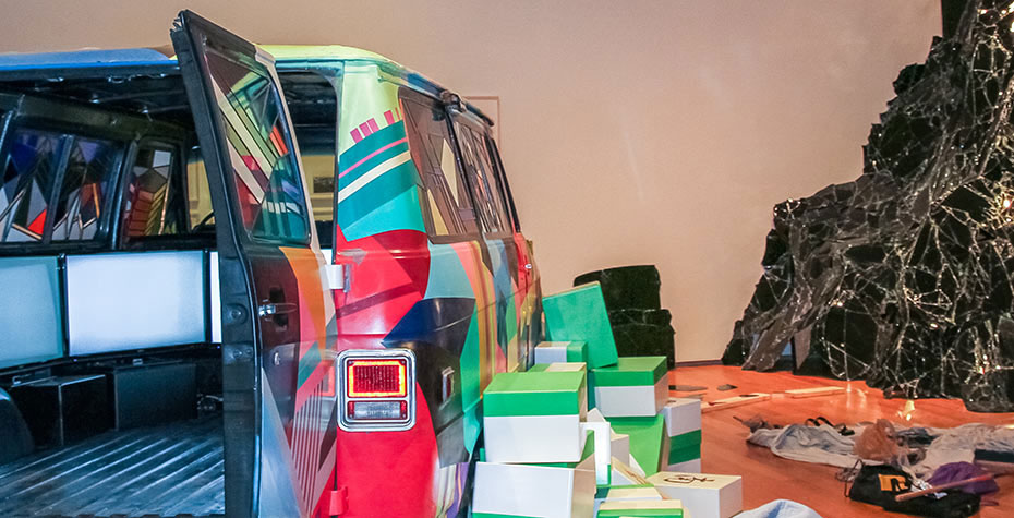 multicolored van with backdoors open revealing may screens inside, boxes and broken glass in installation to the right