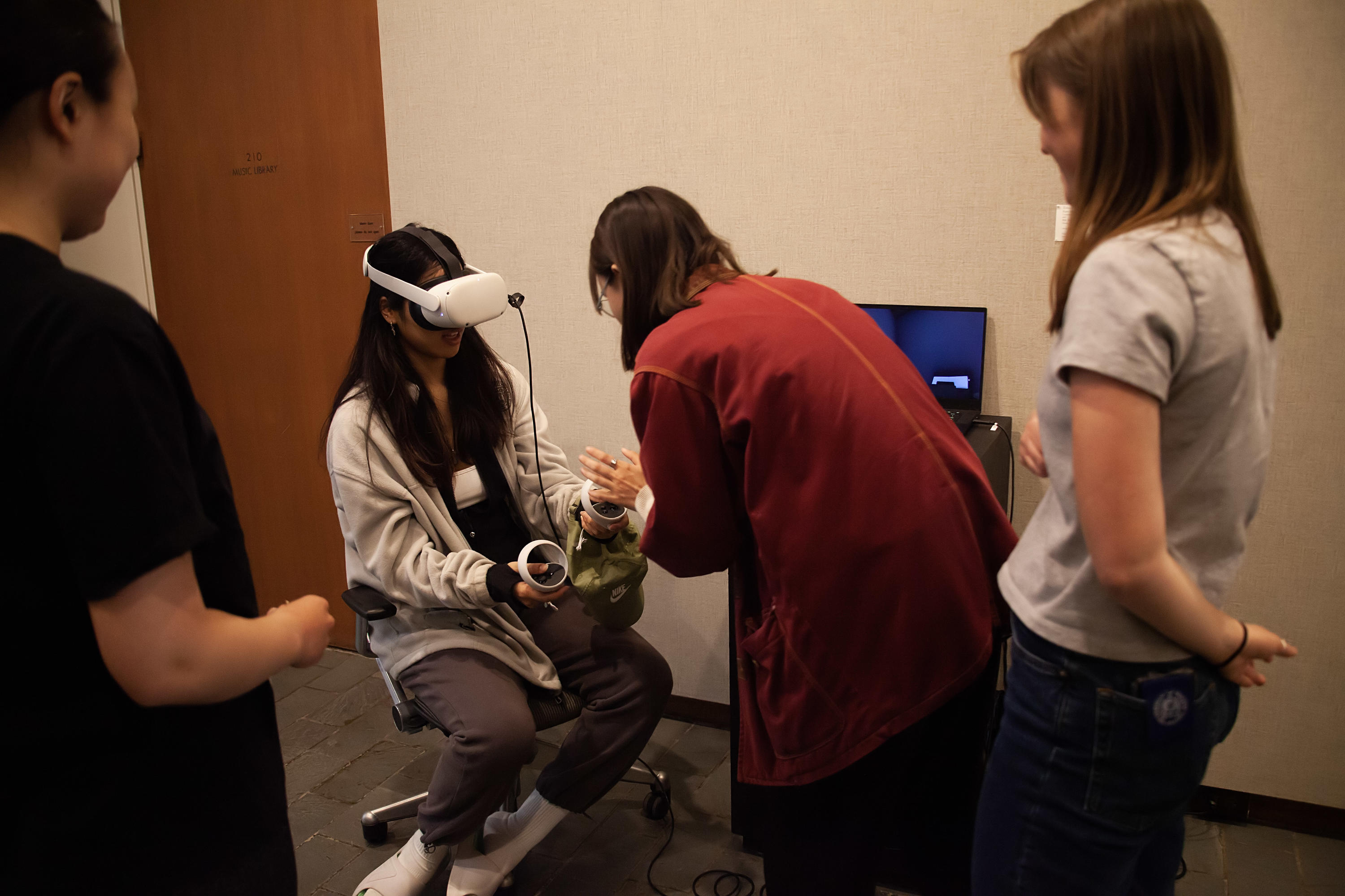 person with long hair sitting in a chair, wearing a white VR headset and holding VR control sticks in their hands, while a person in a red shirt leans over them to explain how the project works. Other people stand at the sides watching.