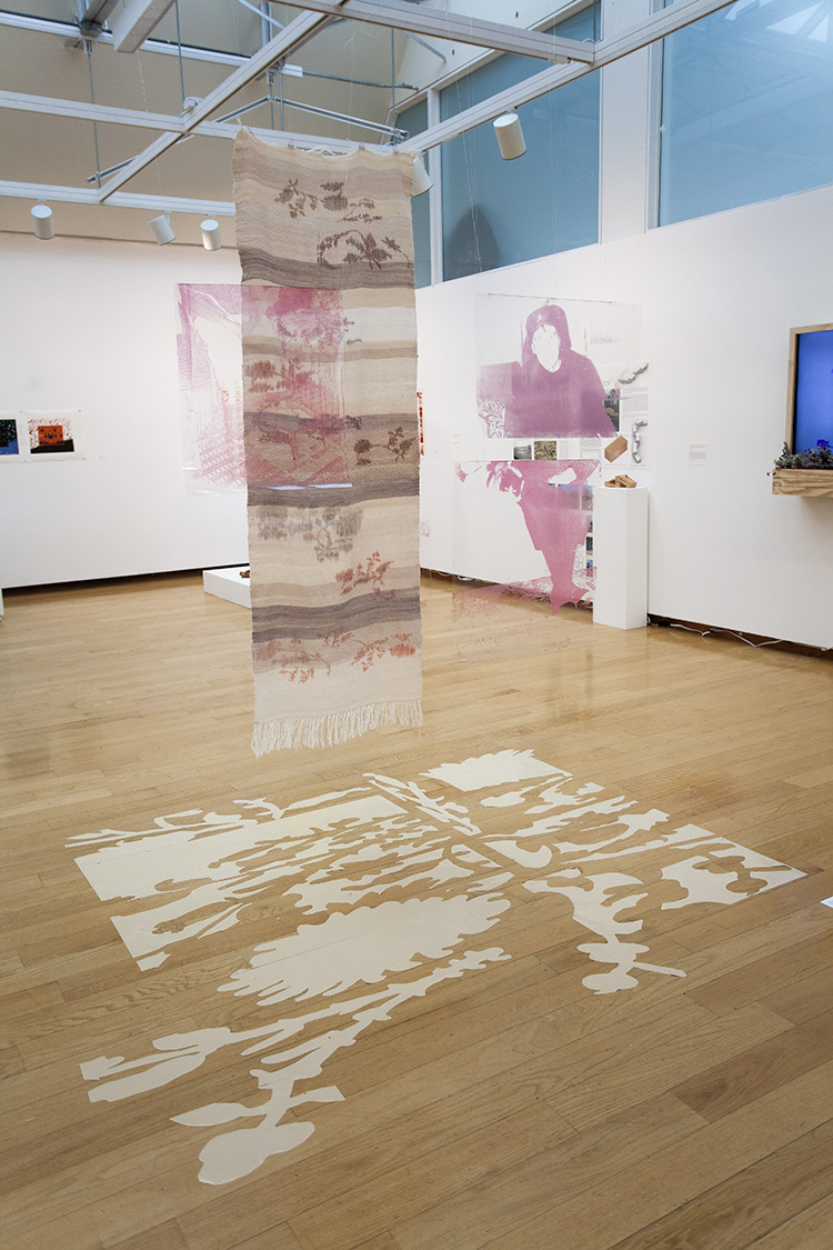 a hanging purple and cream striped textile with reddish floral patterning; also hanging are several transparencies with pink printed overlay; on the floor is a cream warped floral pattern cut out of vinyl