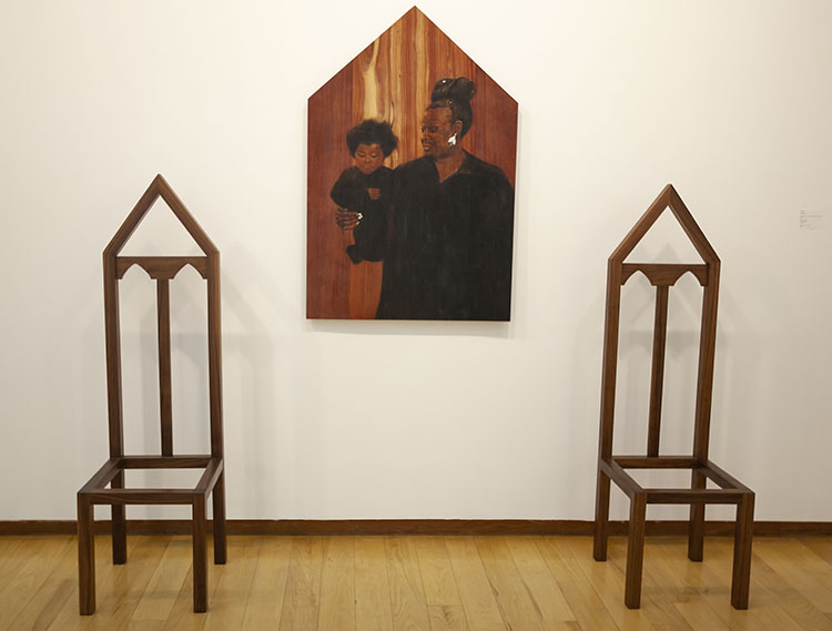 two pointed wooden chairs on either side of a pentagon-shaped wooden panel with pyrography drawing of a woman holding a child
