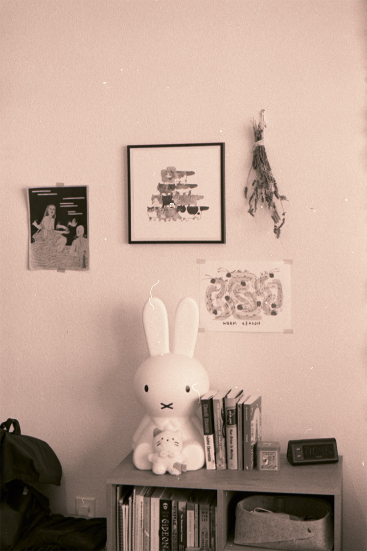 warm-toned photograph of a room with a large miffy the rabbit sculpture, a small cat plush, books, and a clock on a table, prints and some dried plants hanging on the wall