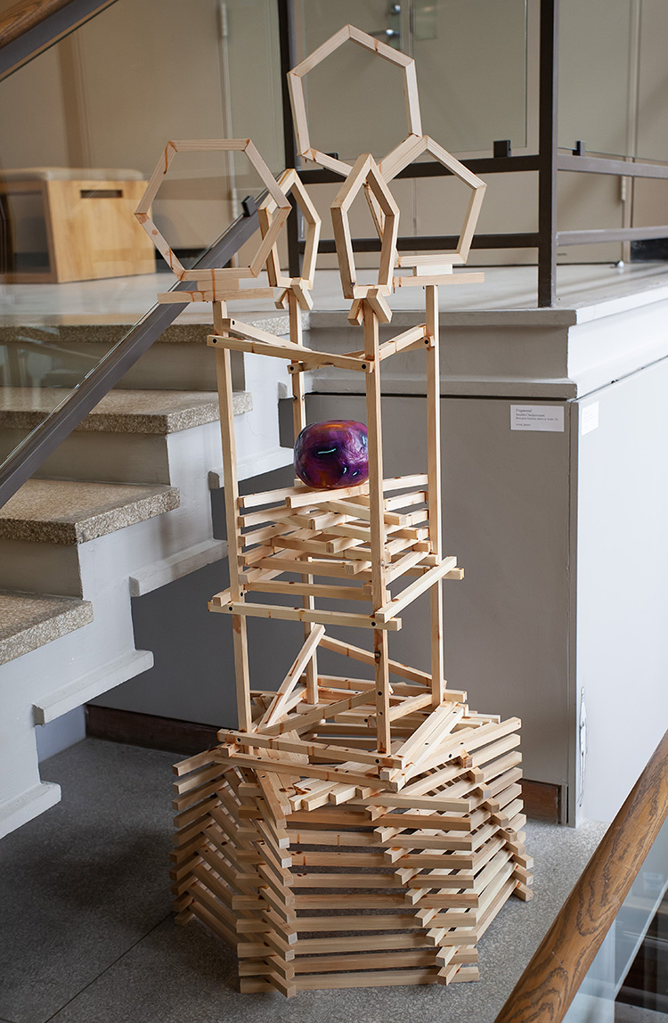 a tall sculpture made of wood 1x1 square rods, with pentagon shapes at top, a stacked wood shelf  with a colorful plaster form on it in the middle, and a pentagonal stacked base