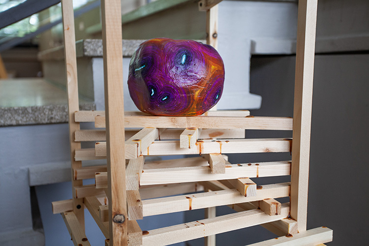 close up on a round plaster form painted mostly in magenta and purple, with thin white and pale blue spots, resting on a wooden structure made up of 1x1 rods