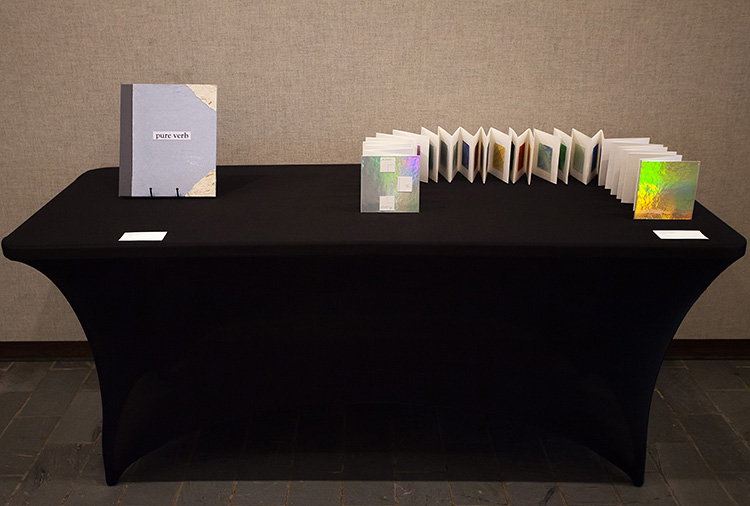 a black cloth-covered table; at left is a book upright on a stand with blue cover and words 'pure verb'; at right is a large accordion book spread out with holographic covers facing the viewer