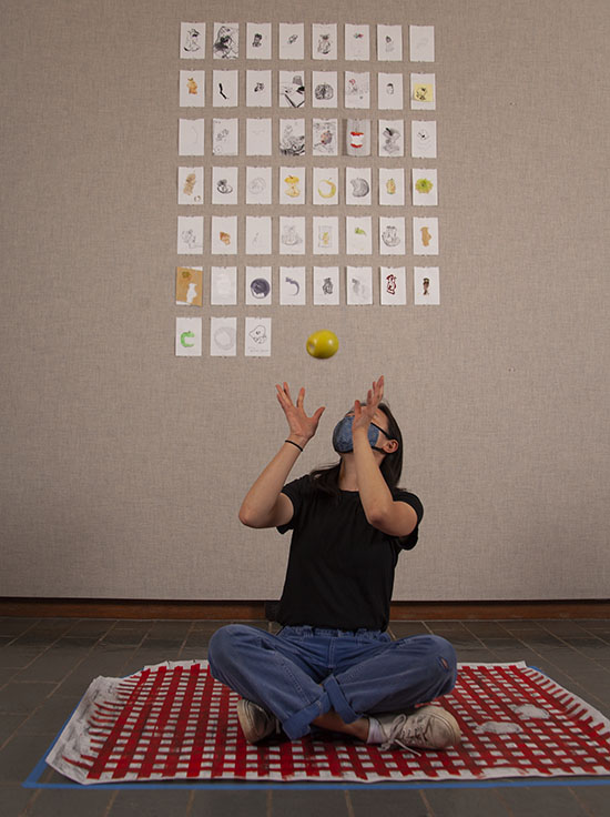 a large grid of small paintings and drawings on a gray fabric wall. In front of them a person in a black tshirt, blue jeans, and blue facemask sits cross-legged on top of a red and white checked piece of paper on the floor. Their hands are up to catch a green apple in midair.
