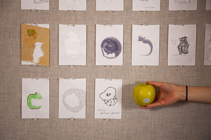 close-up on multiple small drawings hung in rows on a fabric wall. The bottom right drawing's place is taken by a real hand holding a green apple