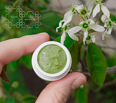 small white pot of green cream, held between two fingers with green and white flower background