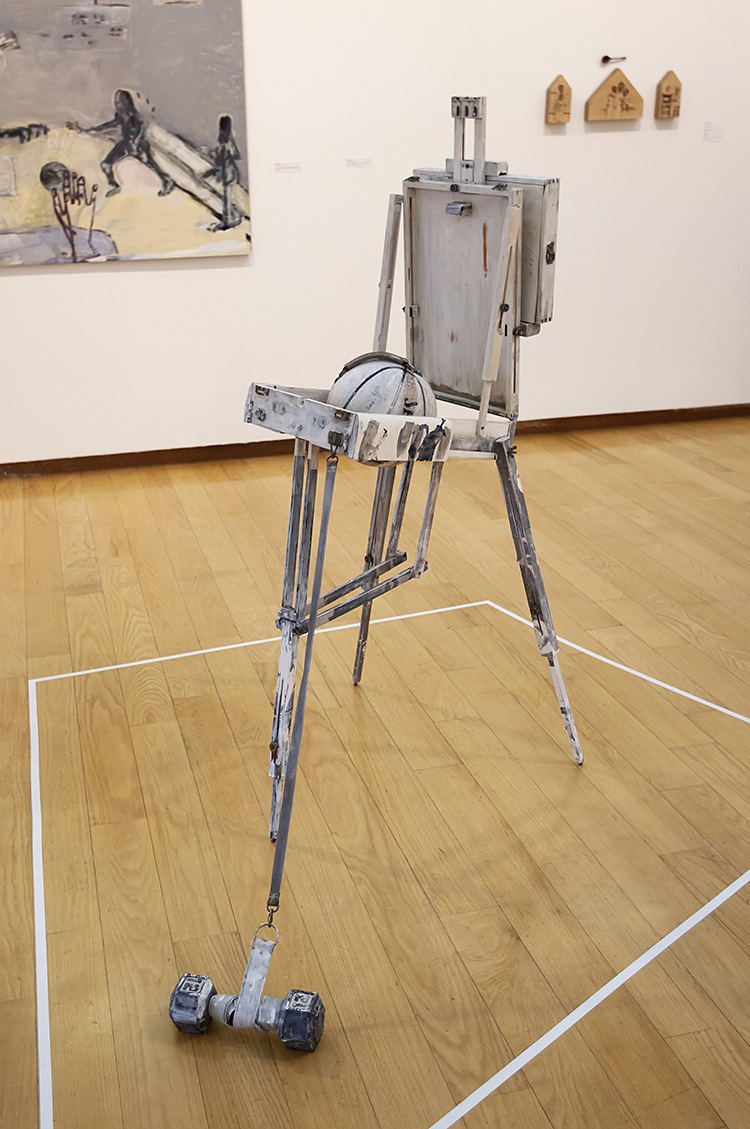 a roughly white painted tripod travelling easel with a hand weight attached to a strap leading from it to the ground, and a painted basketball strapped inside it