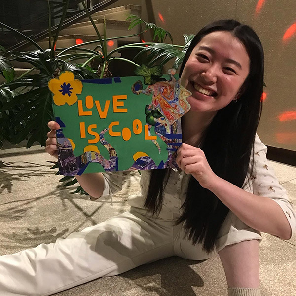 student sitting on floor smiling, holding green paper with collaged images and orange 'LOVE IS COOL' text
