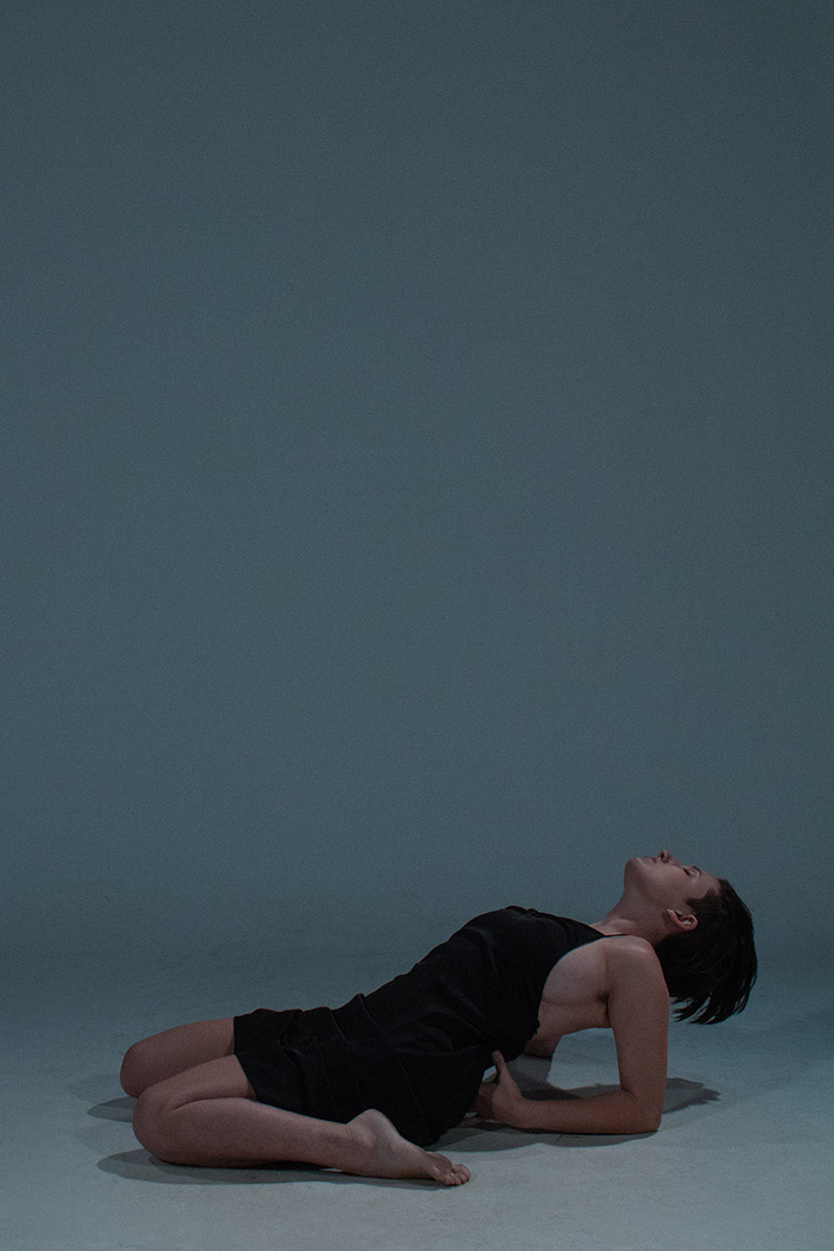 video still of a person in a black dress kneeling on a bluish gray backdrop, leaning back on their elbows with their head thrown back