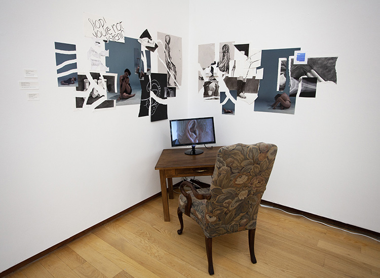 installation in gallery corner of a tilted wood desk with small video screen on it, showing close up of an ear; floral upholstered chair at the desk; walls above covered in collage of ripped papers, drawings, and photographs