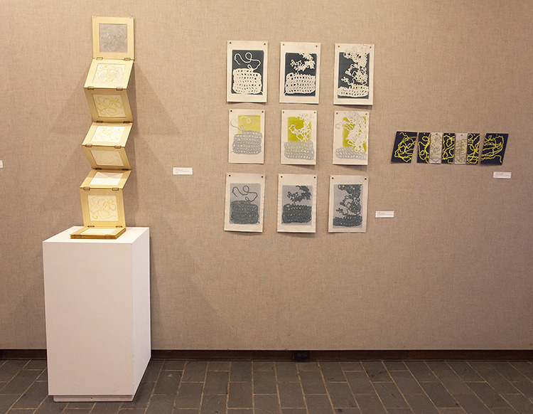 install against a gray fabric wall with long accordion book partially opened against the wall at left, 9-print grid install in center, small horizontal print install with blue and yellow yarn design at right 