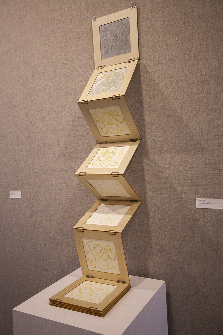 large wooden accordion book with white and yellow prints of string in each page, partially open against a gray fabric wall, piled onto pedestal at the base