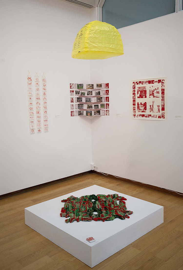 installation in corner of a gallery with yellow medical waste bag lamp above, largely red and white prints on the walls, and an irregular green and red rug on a low pedestal below