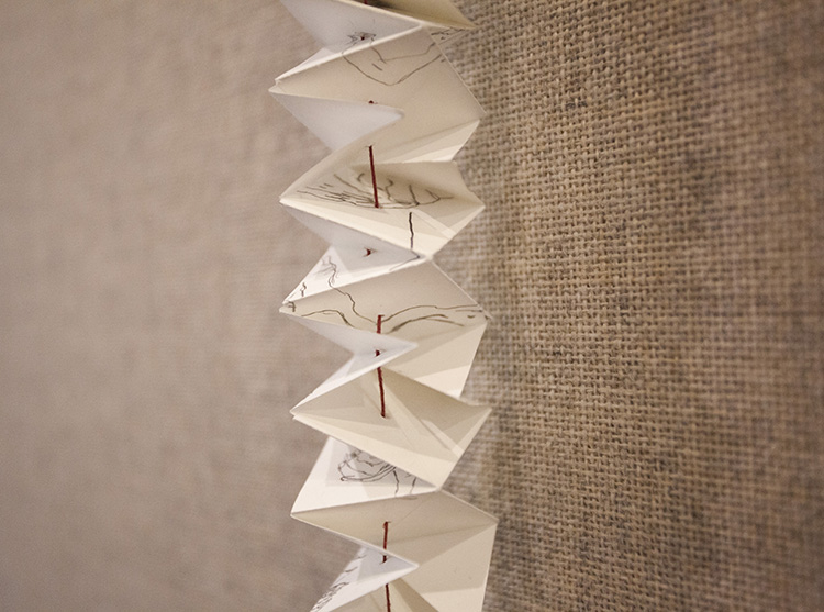 close up on accordioned paper hanging on a thin red thread; the paper has graphite drawings of body parts on it, variably visible