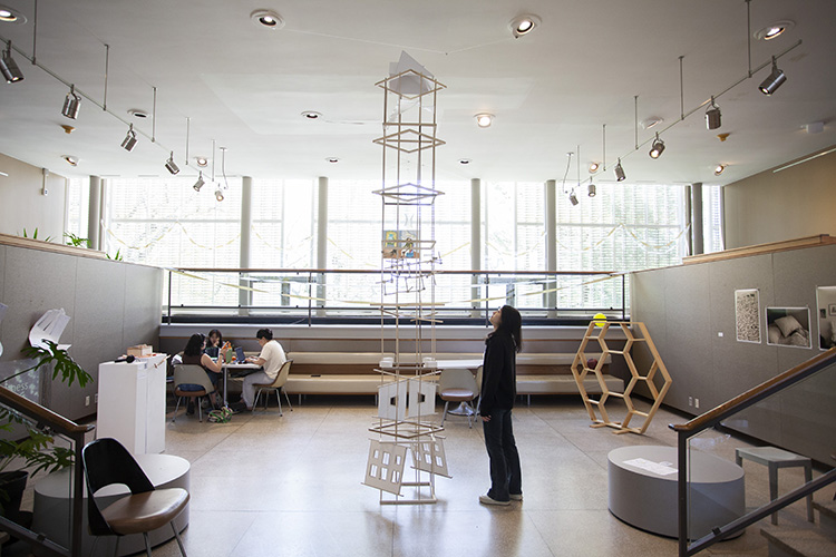 a tall spindly structure made of wood dowels, with objects attached to it at various levels, in a large open space with big windows beyond; a person in dark clothing stands at the foot of the sculpture, craning back to look at it