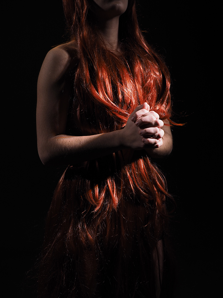 person on a black background, long red hair covering most of their body, hands clasped in front