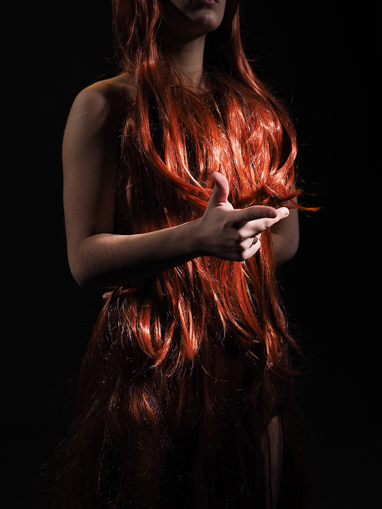 person standing on black background, long red hair mostly covering their body, one hand in front gesturing with thumb and forefinger out