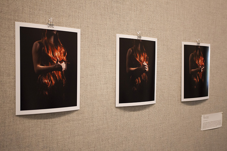 three photographs of a person with long red hair making hand gestures on a black background, installed on a gray fabric wall, viewed from a slight angle