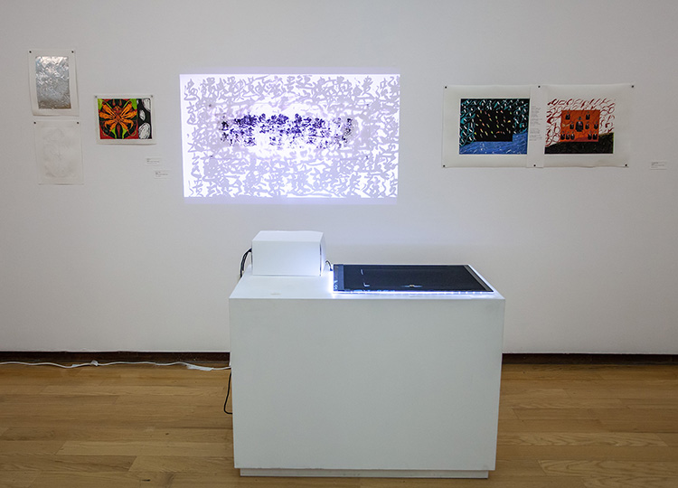 gallery install of 3 prints at left, a video projection of layered chinese text over text print at center, two more prints at right; a pedestal in front of the wall holds a small white box and a flat black light panel