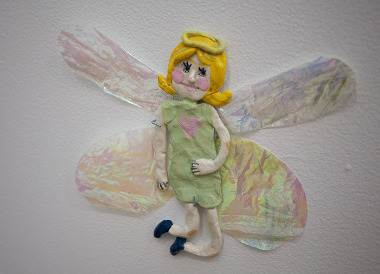 clay fairy/angel sculpture with iridescent wings, green dress with pink heart, blonde hair, and green halo mounted on the wall