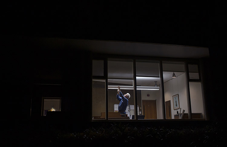 person replacing a long classroom lightbulb, seen from outside a dark building