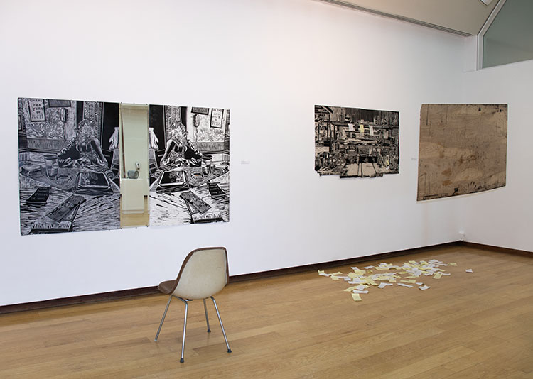 at left, two large black and white woodcuts on either side of a narrow mirror, with a chair in front of them; at right, one large black and white woodcut with a wood board hanging across from it, yellow and white receipts on the floor below it 