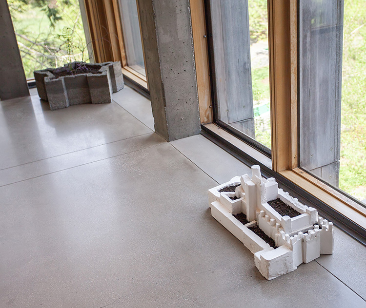 corner of a hallway with gray polished concrete floor and floor to ceiling windows; at left is a gray concrete star shaped form, at right is a white fort model