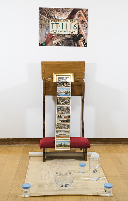 photo of two hands in a chapel surrounding a Michigan 'Water Wonderland' license plate mounted on the wall; below, a prayer bench stuffed with ephemera and a map on the floor with various containers of water, including sample jars