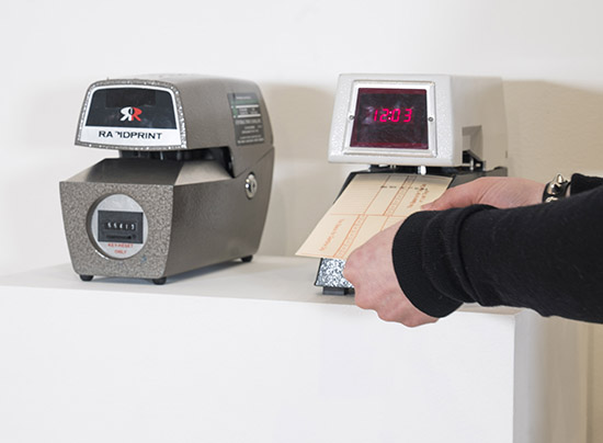 two old fashioned punch clock machines on a pedestal; hands insert a punch card into the machine on the right