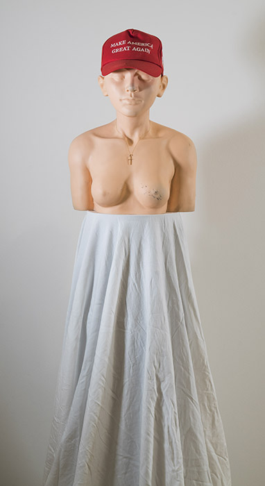 a chest up mannequin with peach skin, breasts, and androgynous face, wearing a red MAGA hat and gold cross chain, on a high pedestal draped with white fabric