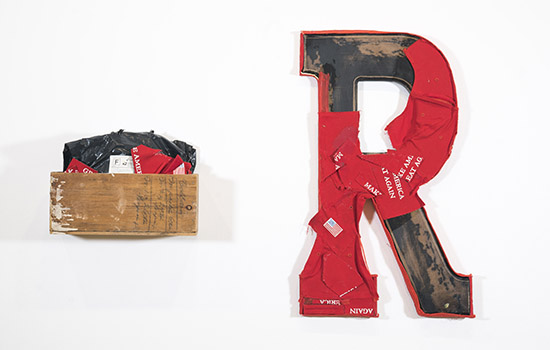 at left, a wood box mounted on the wall with material stuffed inside; at right, a large decorative R mounted on the wall, partially covered in disassembled red hats