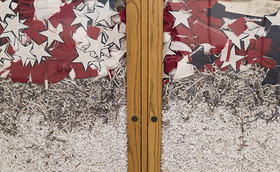close up on glass doors, behind which are tightly squished a shredded American flag and shredded documents