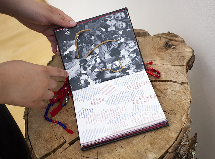 hands holding an artist book with black and white photo collage and attached sticks at left, blue and red text at right, resting on a log