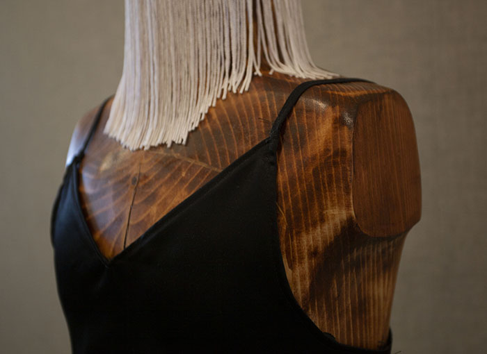 close-up on the shoulder of a stained and carved wood dress form, wearing a black slip. The head of the form has long white fringe draped down its front.