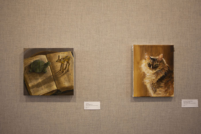 two small paintings on a grayish fabric wall with a label for each; the painting at left shows a toy turtle and gold deer standing on the pages of a book; the painting at right shows a fluffy tabby cat lit dramatically from behind