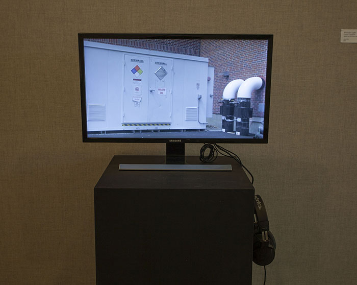 photo of a TV on a black pedestal in front of a grayish fabric wall. The TV screen shows a shot of pipes and a large electrical cabinet on the outside of a brick building