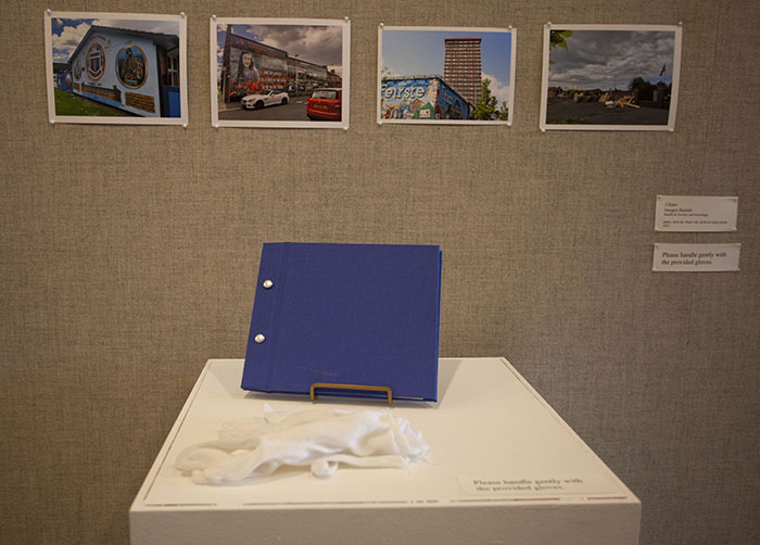 A small blue book on a stand  and white fabric gloves on a white pedestal. The wall behind is gray fabric, and there are 4 color photographs of Belfast locations pinned to the wall above the book.