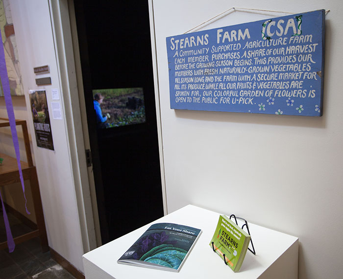 photo of a blue sign with handpainted white text titled 'STEARNS FARM [CSA]'. Below it is a pedestal with a cookbook with lettuce on the cover, and a stack of green postcards for the farm. Beyond them to the left is a dark doorway through which the film screening on a TV can be seen.