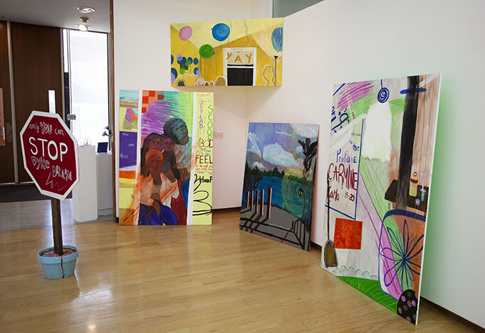 an installation of brightly colored paintings leaned against walls and installed in a corner. On the floor in front of them is a stop sign in a flower pot that reads 'only YOU can STOP DYKE DRAMA'