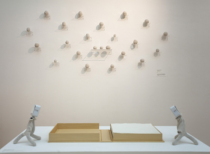 a pedestal with a book in a gold enclosure open on it; on the wall behind, many small balls of yarn are displayed on clear shelves