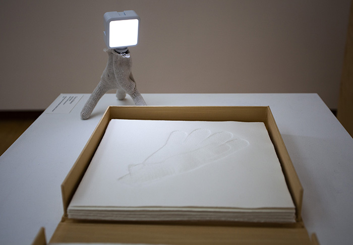 a gold book enclosure with paper embossed with the print of a glove in it. A small light on a tripod wrapped in a glove is behind the book