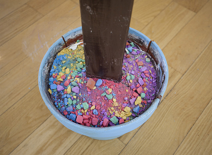 looking down into a light blue cracked flower pot with a brown post in it. Where dirt would normally be, there is instead multicolored ground and broken chalk