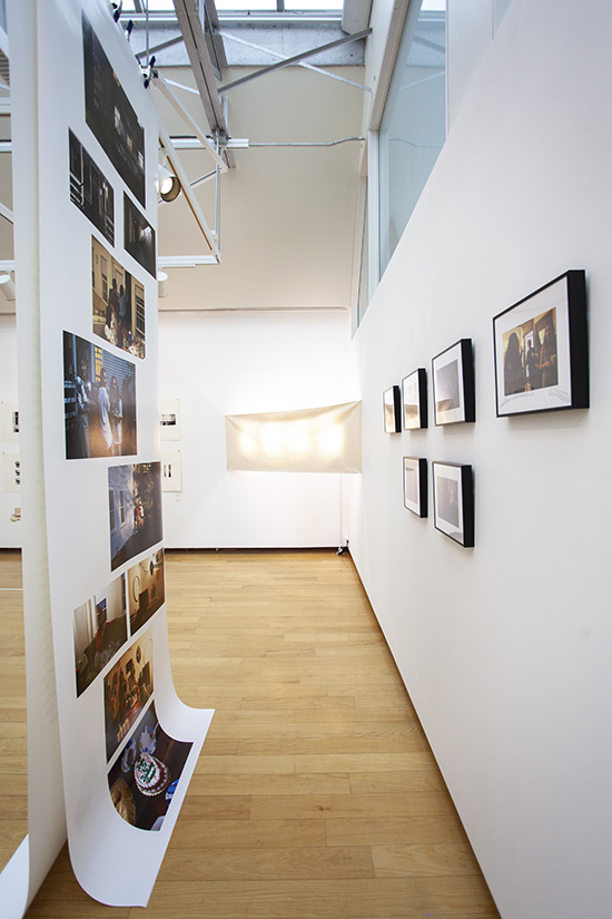 a gallery install with a large hanging piece of paper on which many photos are printed facing a white wall with framed photographs installed on it