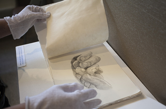 gloved hands turning pages in a handmade book; the visible page shows a graphite drawing of entwined fingers