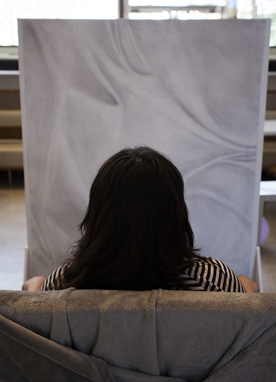 a person with dark hair is seen from behind as they sit in front of a large pencil drawing of draped fabric