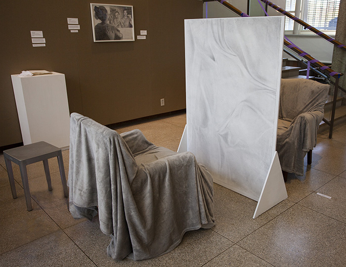 a photo showing an installation of a free-standing graphite drawing of draped paper with chairs covered in fuzzy gray blankets on either side; a white pedestal behind them with a small book on top; a white-framed graphite drawing of two figures on the tan fabric wall behind them
