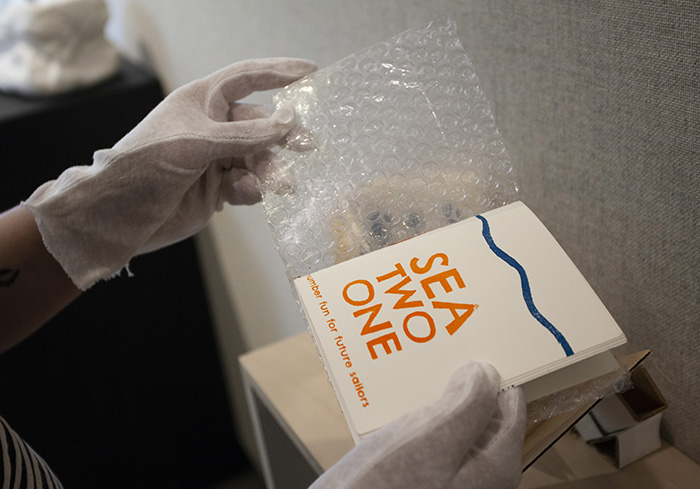 gloved hands holding open a hand-made book. The left page is a cover made of bubble wrap. The right page has a blue squiggly line across the top and reads 'SEA TWO ONE: number fun for future sailors' in orange below the line