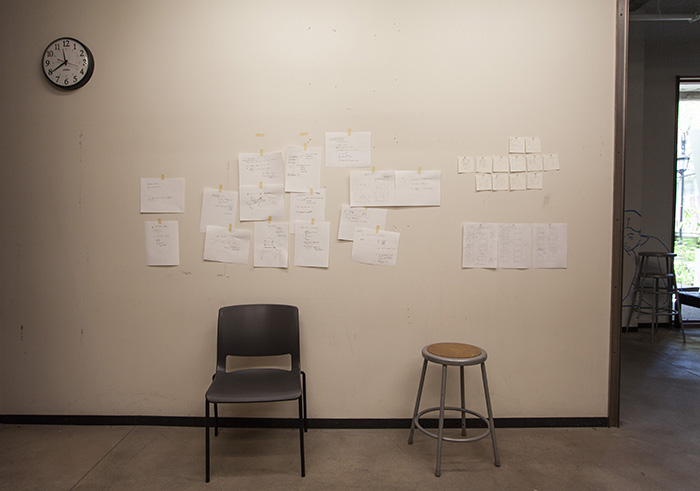 a white wall with a clock at top left, a stool at bottom right, and a black chair roughly center. Many pages of rough notes are taped haphazardly to the wall